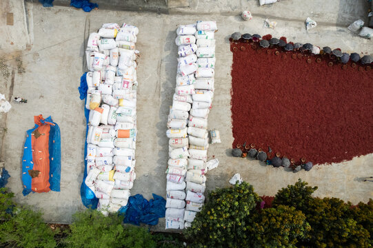 Bogra District, Bangladesh - 14 April 2025: Aerial view of workers drying red chilies on a concrete surface, with rows of stacked sacks and umbrellas providing shade.