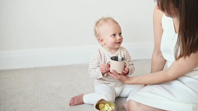 One year old toddler girl learning to drink independently from a silicone training cup while sitting on the floor with her mother in a bright minimalist home with natural daylight.