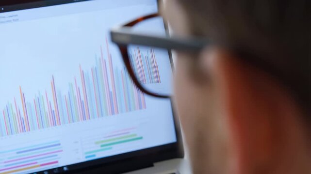Over shoulder view of person wearing glasses reviewing colorful bar charts on laptop screen in office workspace with focused attention and soft ambient light, with glass, laptop, screen, chart detail