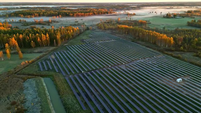 Aerial view of solar farm with rows of photovoltaic panels in a rural landscape surrounded by autumn trees and morning mist in Riga, Latvia.