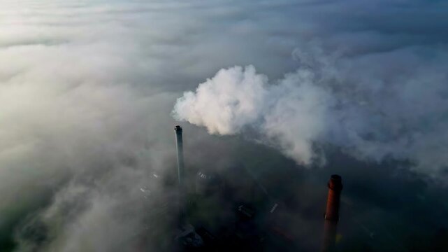 Aerial view of industrial chimneys emitting thick smoke above a dense layer of low-hanging clouds in Riga, Latvia.