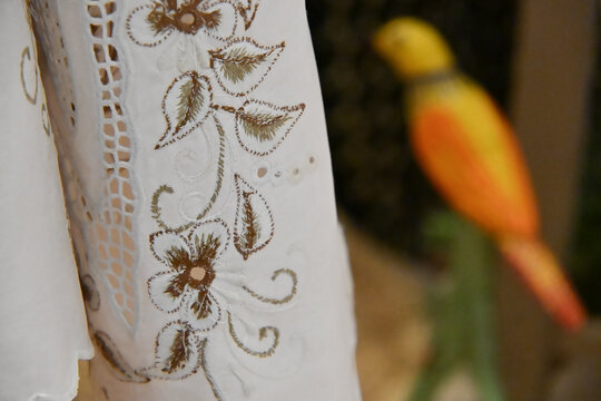 handcrafted white embroidered fabric close up with gold floral embroidery, artisanal lace and leaf pattern detail with blurred wooden bird in background, elegant brazilian embroidery craft display wit