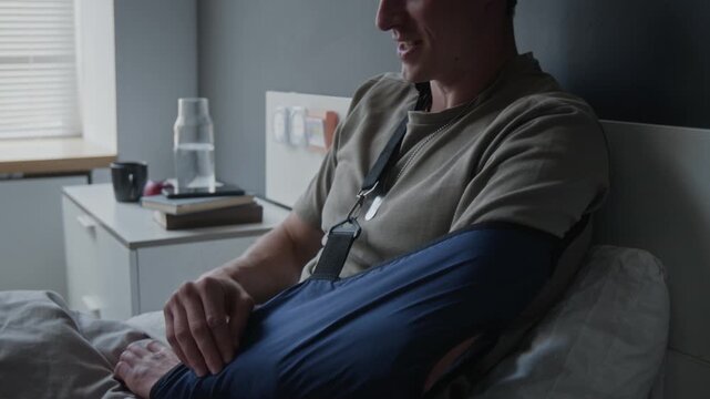 Young Caucasian male patient wearing arm sling lying in hospital bed and discussing recovery with doctor. Ideal for medical insurance, healthcare services, or clinical care advertising