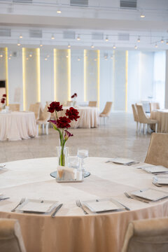 Elegant wedding reception table features a floral centerpiece with red flowers, surrounded by neatly arranged plates and glasses in a bright banquet hall