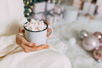 Obraz premium Woman holding cup of cocoa with marshmallow by Christmas tree. Cozy home atmosphere during winter holiday. Warm seasonal beverage for celebration and relaxation. Festive mood winter interior.