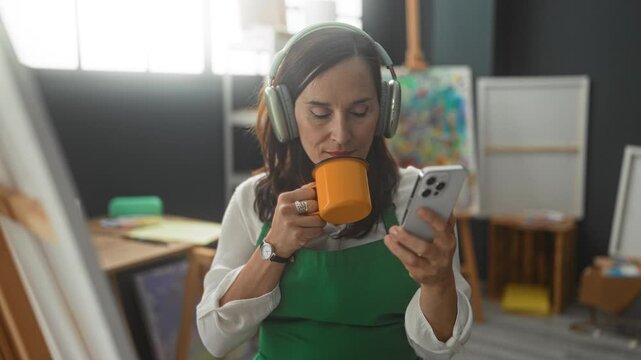 Woman painter in green apron wearing headphones holds orange mug and scrolls smartphone while sipping coffee and smiling in studio; concentration.