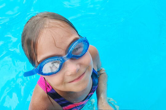 Happy little girl wearing blue swimming goggles in a crystal clear outdoor pool. Summer vacation, active childhood, water fun and healthy lifestyle with copy space.