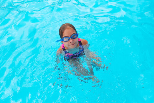 A happy little girl in blue swimming goggles swims in a clear, blue pool. Summer holidays, active childhood, water activities, and a healthy lifestyle.