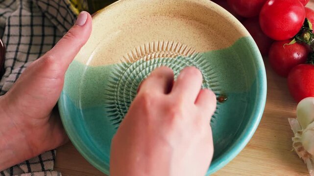 Close-up of hands grating a ripe tomato into a ceramic bowl during food preparation. Fresh tomatoes, garlic and citrus surround the workspace, highlighting authentic Mediterranean cuisine. High