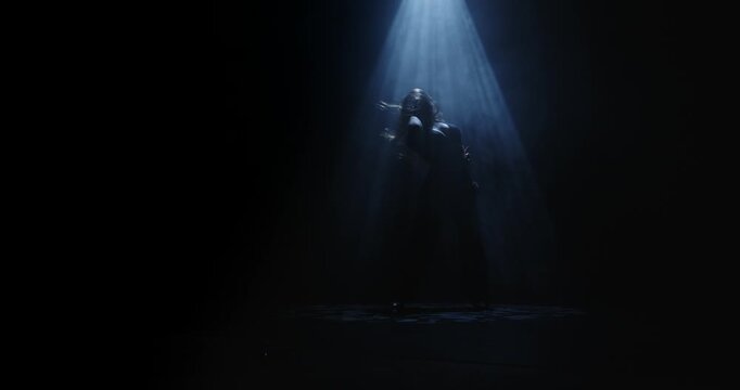 Silhouette of a female dancer performing under a single dramatic overhead spotlight on a dark stage. Concept of dance, performance, nightlife and art, fitness, fashion and entertainment advertising.