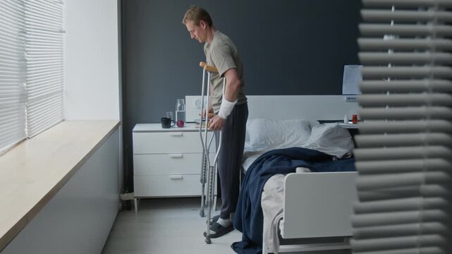 Young adult Caucasian man with bandaged arm standing up from hospital bed and walking with crutches. Clip for medical insurance, rehabilitation services, or healthcare marketing