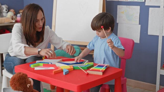 Woman guides boy's hand to hold a mallet and taps colorful xylophone and wooden blocks on a red table in a building; learning bonding.