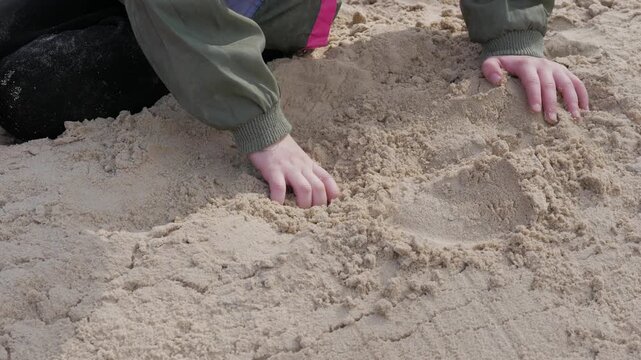 A child wearing a jacket digs hands into soft beach sand outdoors during daytime play.