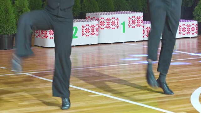 Two performers in dark trousers synchronize dynamic leg movements during a dance routine on a polished wooden parquet floor.