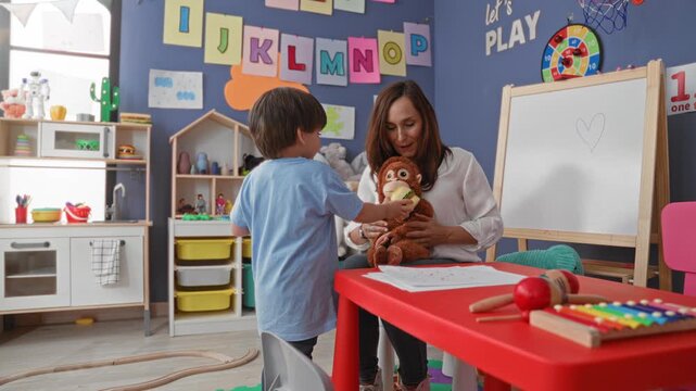 Woman teacher holds stuffed monkey while watching little boy play at toy kitchen in preschool classroom; nurturing care.
