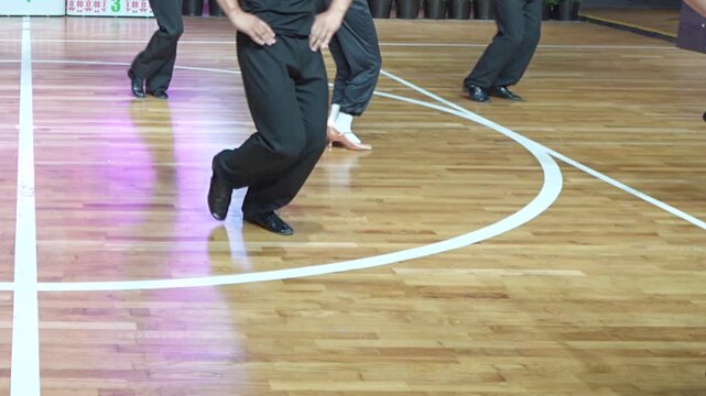 A group of performers dressed in black trousers and dance shoes executes synchronized footwork on a polished wooden parquet floor.