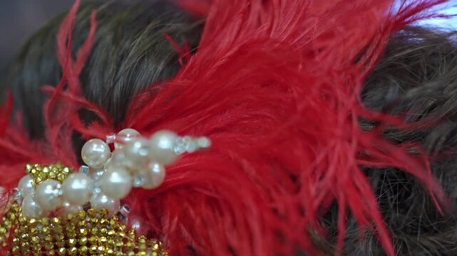 A vintage roaring twenties style headband adorned with white pearls, gold rhinestones, and vibrant red feathers rests in dark hair.