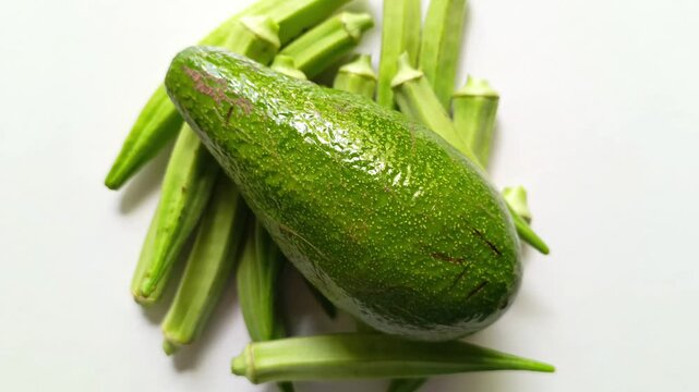 Whole fresh raw avocado Persea americana resting on a pile of green okra Abelmoschus esculentus pods isolated on clean white background in Guatemala