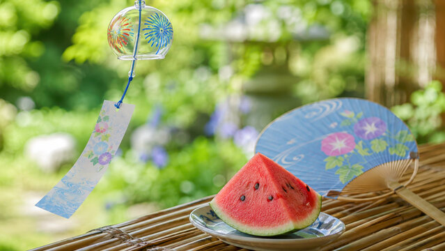 Glass wind chime with painted fireworks, paper fan near watermelon slice on bamboo porch, green garden bokeh, japanese summer atmosphere, obon seasonal refreshment, nostalgic cooling breeze