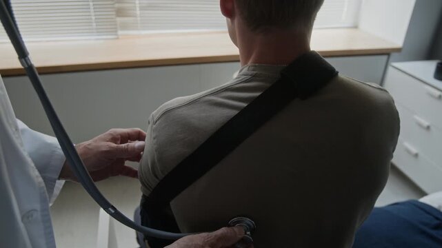 Mature male doctor using stethoscope to examine back of young adult patient wearing shoulder brace. Ideal for healthcare services, medical insurance, or injury recovery ads