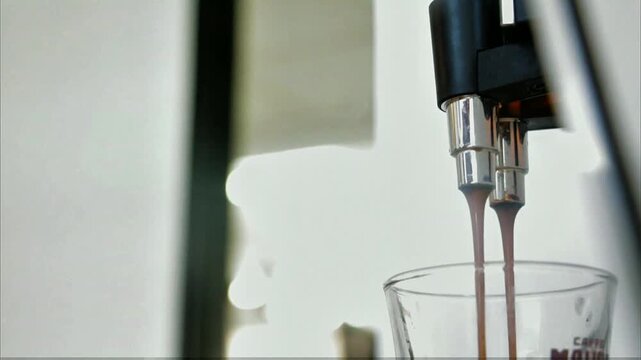 Coffee machine dual metal spouts positioned over clear glass cup, bright soft background with gentle blurred shapes