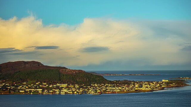View of coastal town nestled along the shoreline with rolling hills and calm sea under a cloudy sky in Alesund, More og Romsdal, Norway.