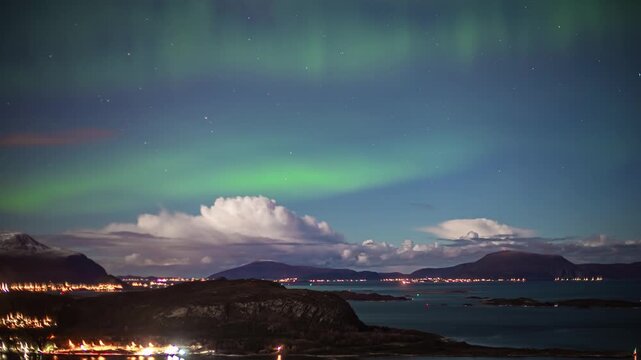 View of vibrant green aurora borealis illuminating the night sky above the coastal mountains and city lights of Alesund, More og Romsdal, Norway.