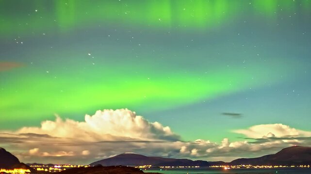 View of the vibrant green Aurora Borealis illuminating the night sky above the mountains and distant city lights of Alesund, More og Romsdal, Norway.