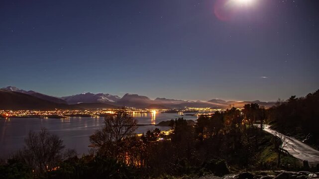 View of night cityscape of Alesund with illuminated buildings reflecting on the calm fjord waters under a starry sky with distant mountains, Alesund, More og Romsdal, Norway.