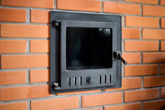 Cast iron stove door with glass built into brick wall of traditional sauna. Metal door features air vents and handle for safe wood firebox regulation inside heating room.