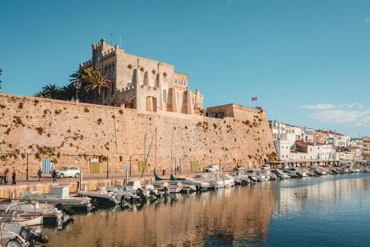 View of the historic fortifications and buildings overlooking the harbor with moored boats in the calm water under a clear blue sky in Ciutadella, Balearic Islands, Spain.