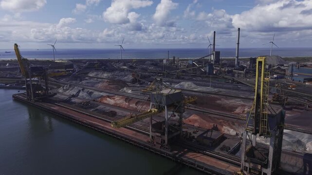 View of the Tata Steel industrial complex featuring large stockpiles of raw materials, industrial cranes, and wind turbines along the coast in Velsen-Noord, Noord-Holland, Netherlands.