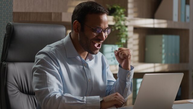 Happy cheerful Indian business man celebrating victory at office laptop winning American executive businessman lucky male excited good news success online bet win achievement computer triumph indoors