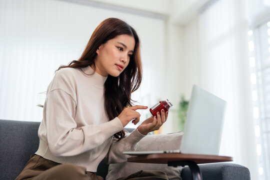 Woman holding pill bottle and using laptop computer searching for information about  prescription medicine while sitting in livingroom at home .
