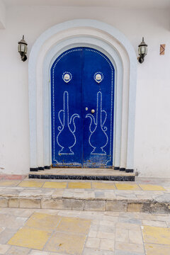 View of a traditional blue arched door with intricate white nail patterns and wall lamps set in a white wall in Kairouan, Kairouan Governorate, Tunisia.