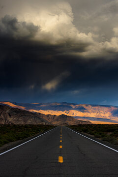 View of Death Valley National Park with a straight road leading towards mountains under dramatic dark storm clouds and golden light in the United States.