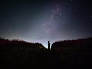 Silhouette of a person standing on a grassy hill against a breathtaking night sky full of stars © Hamid