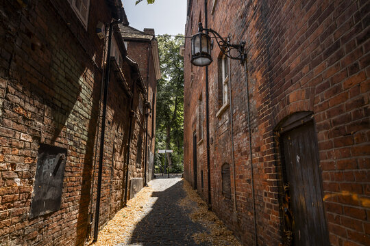 View of a narrow alleyway lined with weathered red brick walls, featuring an old-fashioned street lamp and a cobblestone path covered in fallen leaves, Coventry, England, United Kingdom.
