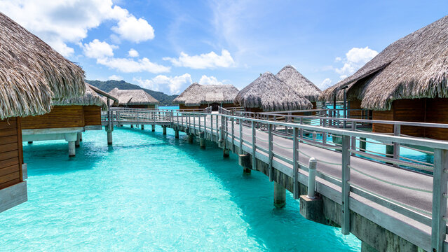 View of overwater bungalows with thatched roofs connected by a wooden walkway over the turquoise lagoon in Bora-Bora, Leeward Islands, French Polynesia.