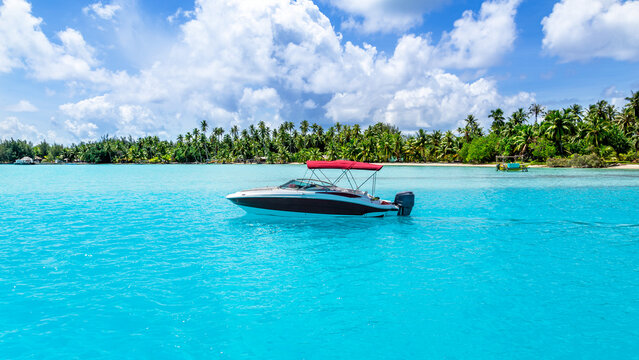 View of a motorboat with a red canopy floating on clear turquoise water with a lush palm-fringed coastline in the background in Bora-Bora, Leeward Islands, French Polynesia.