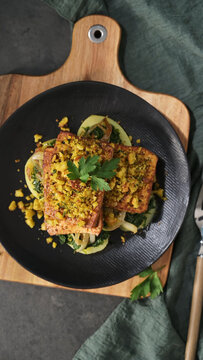 Fried tofu cutlet with breadcrumb crust and vegetable bed served on a dark plate