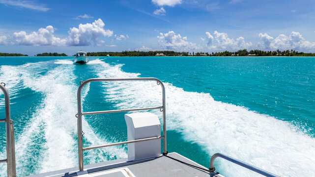 View of a boat wake trailing through vibrant turquoise lagoon waters with a distant boat and tropical coastline under a bright blue sky in Bora-Bora, Leeward Islands, French Polynesia.