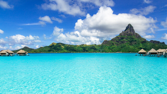 View of overwater bungalows in a turquoise lagoon with Mount Otemanu in the background under a blue sky in Bora-Bora, Leeward Islands, French Polynesia.