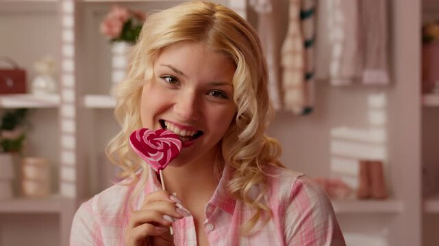 Joyful Caucasian teen girl biting heart shaped lollipop in pink bathroom smiling looking at camera blonde woman lady enjoying sweet candy playful dessert treat glamour beauty carefree inside home bath