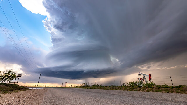 View of a massive supercell storm with dramatic rotating clouds looming over a rural road and an oil pump jack in Oklahoma City, Oklahoma, United States.