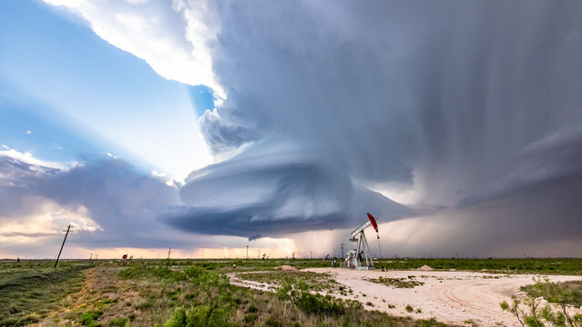 View of a dramatic supercell storm cloud hovering over an oil pumpjack in a rural field with light rays breaking through the clouds in Oklahoma City, Oklahoma, United States.