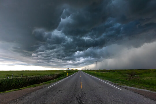 View of a long straight road stretching into the distance under a dramatic and heavy dark storm cloud formation over the green landscape in Oklahoma City, Oklahoma, United States.