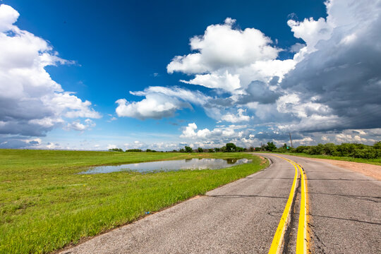 View of a rural road with yellow lane markings curving through a green field with a puddle under a dramatic sky with heavy storm clouds in Oklahoma City, Oklahoma, United States.