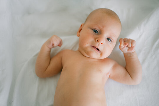Newborn baby boy lying on a white sheet, flexing both arms like a strongman, creating a humorous and adorable portrayal of childhood strength and potential growth