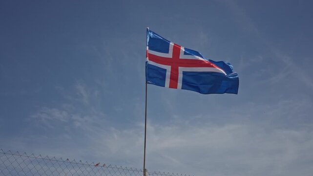 Iceland flag with red cross and white border waving on a tall pole above rusted chainlink fence on street; pride serenity.